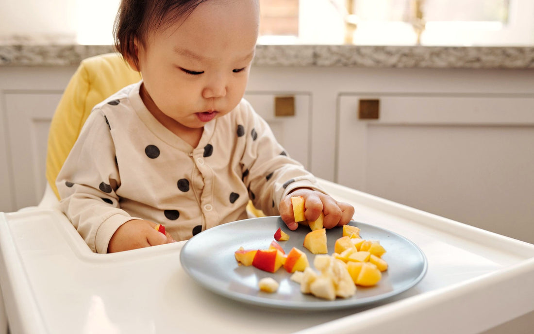 Smiling baby in a high chair finger-feeding fruit from a silicone plate